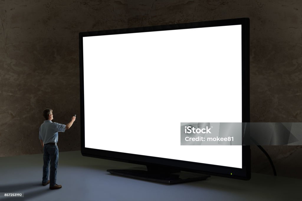 Man pointing tv remote control at world’s biggest giant television with blank screen Man pointing tv remote control at world's biggest television that towers over him shining bright light in a dimly lit room with clean blank screen for your message and image sending a powerful message Large Stock Photo