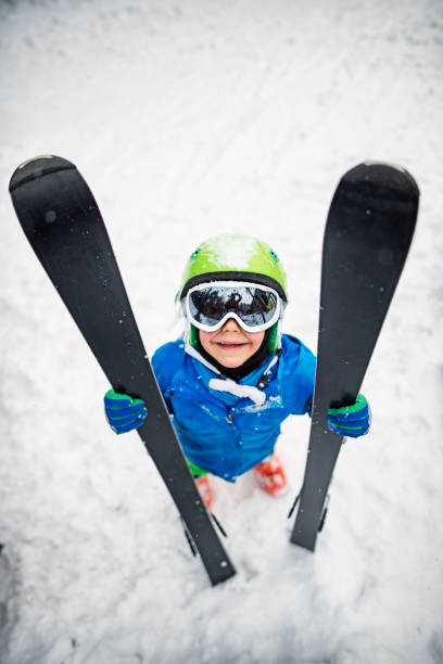 Little boy holding skis and looking up into the camera.\n