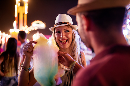 Couple eating cotton candy at amusement park, incidental people in background