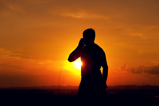 Silhouette of young man talking on the phone, enjoying sunset time outdoors. Travelling and vacation concept.