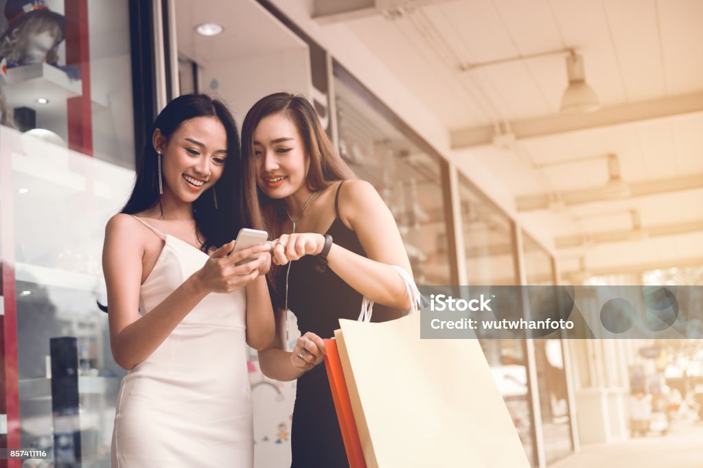 Two asian woman standing at store front with happy watching smart phone at shopping mall center. Shopping Stock Photo