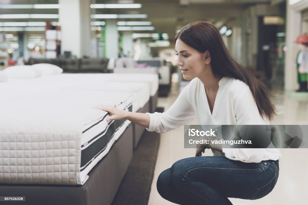 A woman chooses a mattress in a store. She sits next to him and examines him A woman in a white shirt and jeans in a mattress store. She examines the mattress she wants to buy. She squats and looks at the mattress Mattress Stock Photo