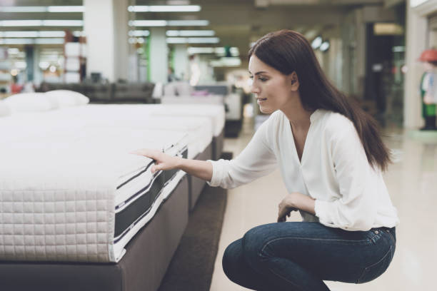 A woman chooses a mattress in a store. She sits next to him and examines him A woman in a white shirt and jeans in a mattress store. She examines the mattress she wants to buy. She squats and looks at the mattress mattress stock pictures, royalty-free photos & images
