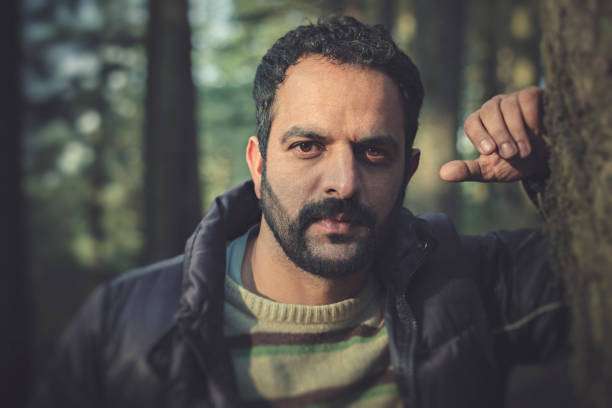 Outdoor image of Asian, Indian young, strong, masculine, man standing near tree trunk and looking at camera with blank expression. He is in warm cloths. One person, waist up and selective focus with copy space. Image shot in himachal Pradesh.