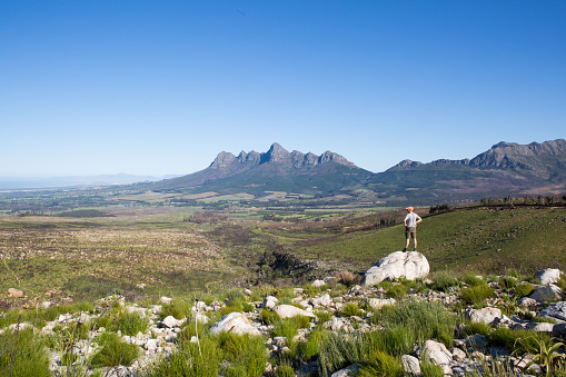 A young male on a hiking adventure stands on a rock with his arms on his hips and admires the view - Helderberg, South Africa