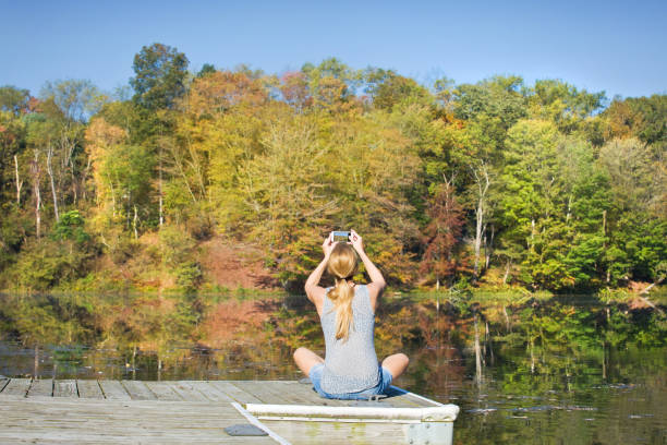 A woman sitting on a boat dock and taking a photo on her phone of the beautiful scene around her.
