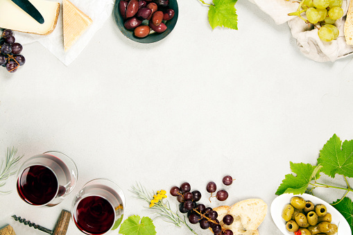 Wine and snack set. Variety of cheese, olives, prosciutto meat, baguette slices, black grapes and glasses of red wine over grey marble background, top view, copy space