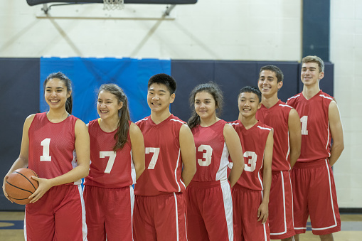 Co-ed, multiethnic high school basketball team poses for a picture. They are lined up, smiling, in red jerseys and shorts, and the female on the left is holding a basketball.