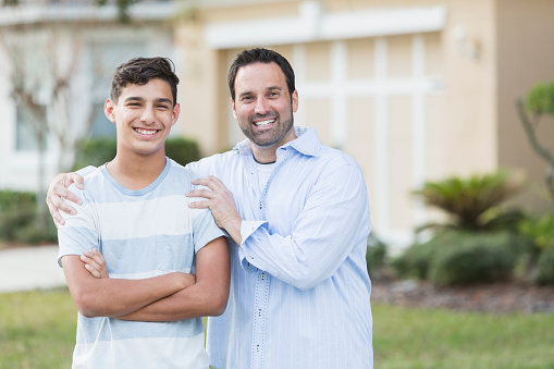 A father and his 13 year old teenage son standing in their front yard, smiling confidently at the camera.