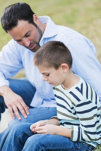 A father with his 7 year old son sitting on the street curb, looking down with serious expressions. The little boy looks sad and upset, and his father is being supportive.