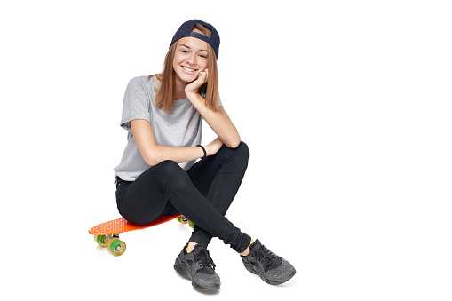 Teen girl in full length sitting on skate board smiling at camera, isolated on white background
