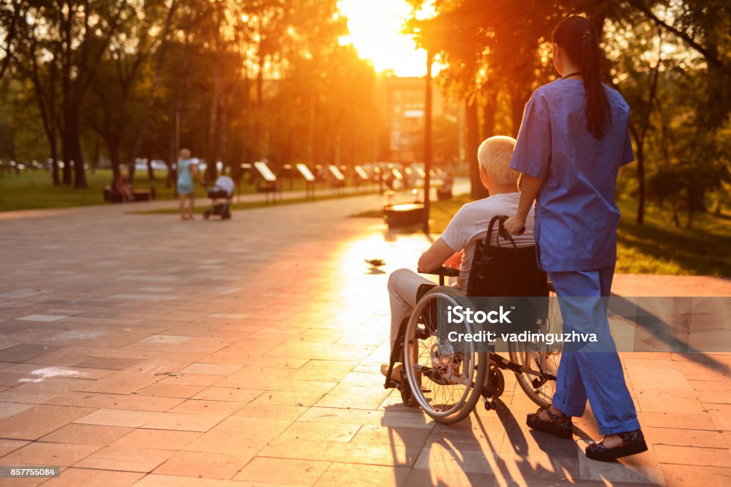 Una enfermera y un anciano que se sienta en una silla de ruedas paseando en el parque al atardecer - Foto de stock de Paciente libre de derechos