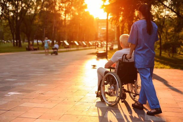A nurse and an old man who sits in a wheelchair strolling in the park at sunset stock photo