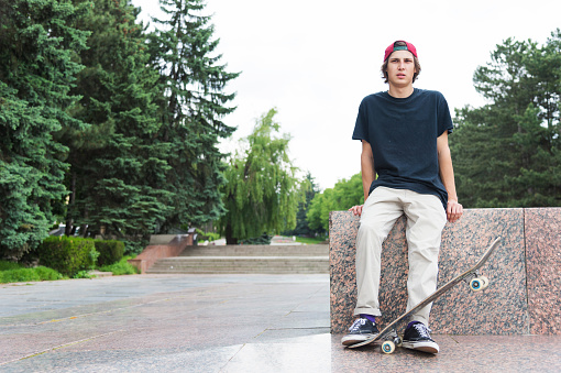 The long-haired skater in a cap and T-shirt sits with his back to the camera and thinks next to the skateboard against the background of the landscape landscape.