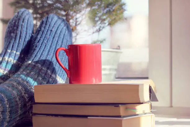 red mug on a pile of books on background feet in warm socks on a table next to the window