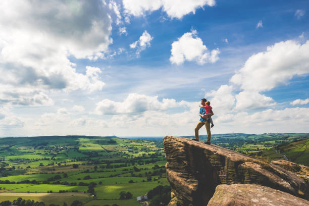 Father and Son Hiking in Peak District A father and his young son stand on a rocky outcropping overlooking the valley below at The Roaches, Peak District National Park, Derbyshire, United Kingdom. The father is spending some quality bonding time with his son and showing him the beauty of England on a summer afternoon. peak district national park stock pictures, royalty-free photos & images