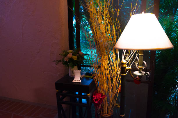 corner of a room with a vintage set consisting of a lamp, two white roses in glass bottles and a glass with a flower on an old wooden table, all lit with natural light only.