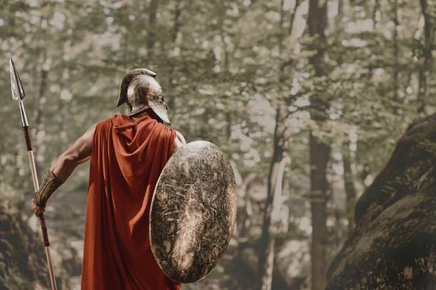 Anonymous warrior in gladiator outfit Back view of man in gladiator helmet and red long cloak standing with round shield and spear in hands looking away in woods. Spartan. armory photos stock pictures, royalty-free photos & images
