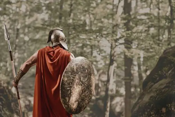 Back view of man in gladiator helmet and red long cloak standing with round shield and spear in hands looking away in woods. Spartan.