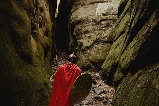 Back view of Roman gladiator wearing red cloak and helmet walking with arms in mossy canyon exploring location. Spartan.