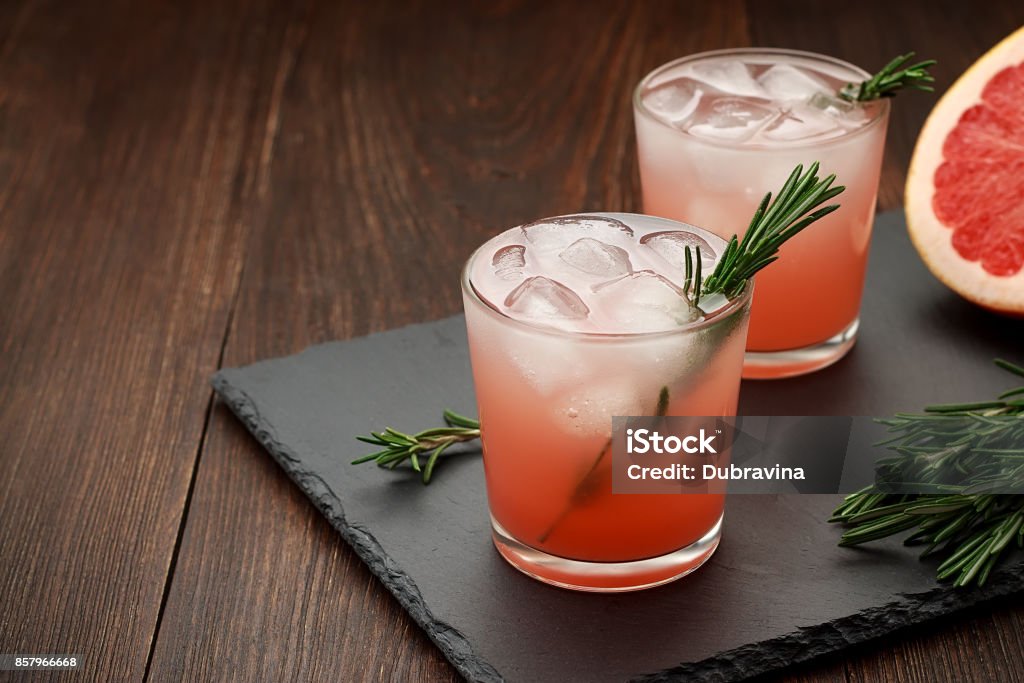 Two glasses of citrus summer drink with grapefruit and rosemary on dark background. Two glasses of citrus summer drink with grapefruit and rosemary on dark background. Detox fruit cocktail with ice. Copy space. Cocktail Stock Photo