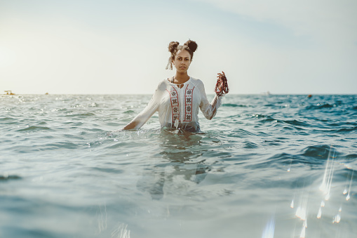 Charming young black curly lady in traditional African chemise standing up to the waist in teal seawater and holding beads in her left hand, strong reflection in the bottom, ripples on water surface