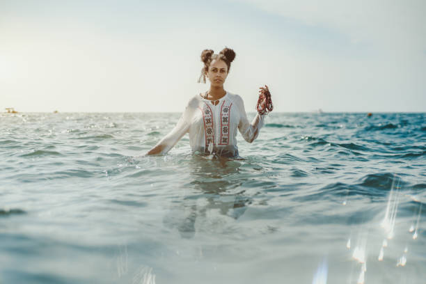 Black girl in teal water Charming young black curly lady in traditional African chemise standing up to the waist in teal seawater and holding beads in her left hand, strong reflection in the bottom, ripples on water surface walking in water stock pictures, royalty-free photos & images