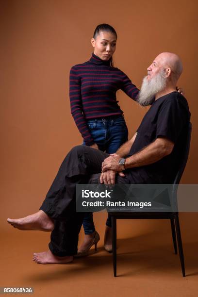 Studio Shot Of Mature Bald Man With Long Beard And Young Beautiful Asian Transgender Woman Against Colored Background Stock Photo - Download Image Now