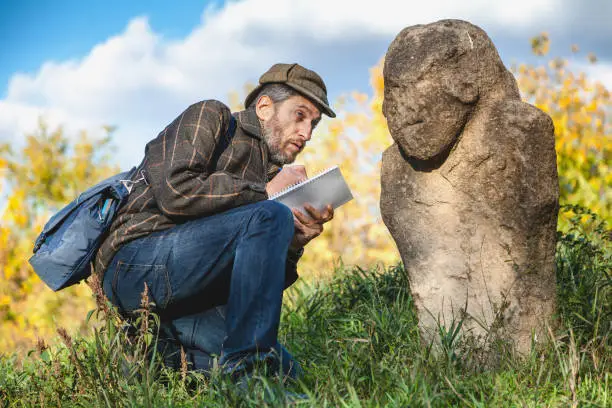 Scientists with a beard in a plaid jacket sits and sees the stone monument of Scythian epoch on a green hillside and makes notes in a notebook