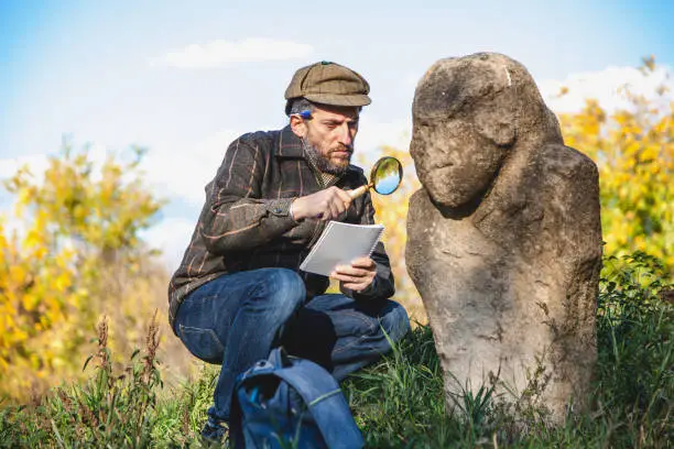 Scientists with a beard in a plaid jacket examines through a magnifying glass stone monument Scythian epoch on a green hillside