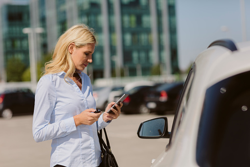 Businesswoman talking on the phone outdoors. Standing in front of the car and typing on her smart phone.