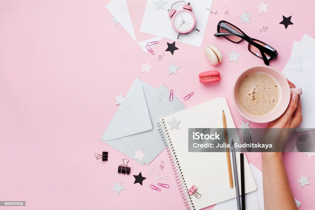 Frau Hand mit Tasse Kaffee, Macaron, Bürobedarf und leeren Notizbuch auf rosa Pastell Tischplatte Ansicht. Mode-Bloggerin Arbeitstisch. - Lizenzfrei Schreibtisch Stock-Foto