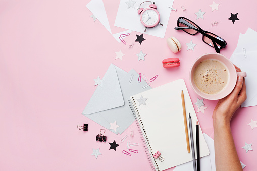 Woman hand with cup of coffee, macaron, office supply and empty notebook on pink pastel table top view. Fashion female blogger working desk.