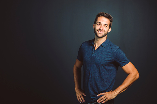 Smiling fashionable male is with hands on hip. Portrait of handsome man is standing against gray background. He is wearing casuals.