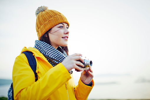 Beautiful young woman looking away while holding old-fashioned camera against clear sky. Female hiker is wearing yellow knit hat and jacket. She is on vacation during winter.