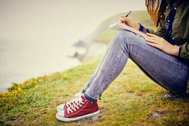 istock Young woman writing in diary on grassy cliff by sea 858148176