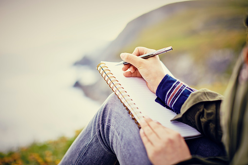 Midsection of young woman writing in diary while sitting on mountain. Cropped image of female tourist spending leisure time in nature. Focus is on pen held by her.