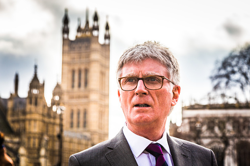 Close up color image depicting a senior businessman, of caucasian ethnicity, standing outside the famous buildings of the Houses of Parliament in Westminster, London, UK. The man is wearing a smart grey suit, white shirt and a purple necktie. He is wearing fashionable spectacles, too. The man has white hair (he is in his 60s) and is looking off past the camera. The famous London architecture is blurred nicely out of focus in the background. Lots of room for copy space.