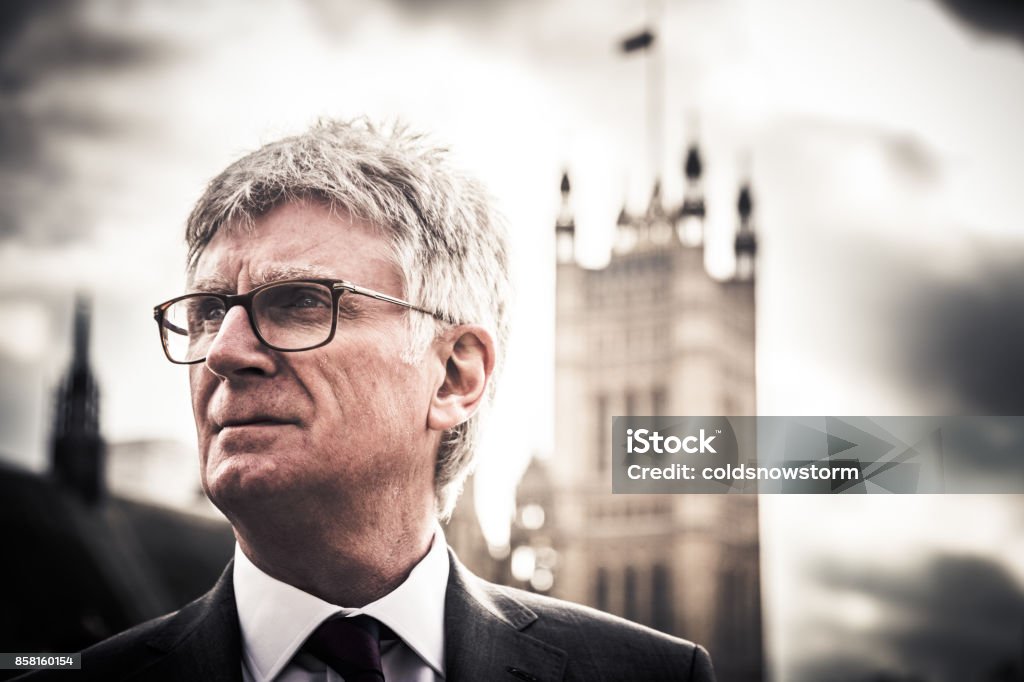 Close up of senior caucasian businessman outside houses of parliament, London, UK Close up color image depicting a senior businessman, of caucasian ethnicity, standing outside the famous buildings of the Houses of Parliament in Westminster, London, UK. The man is wearing a smart grey suit, white shirt and a purple necktie. He is wearing fashionable spectacles, too. The man has white hair (he is in his 60s) and is looking off past the camera. The famous London architecture is blurred nicely out of focus in the background. Lots of room for copy space. Member of Parliament Stock Photo