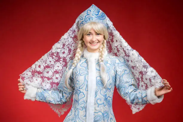 Beautiful girl dressed in Snow Maiden costume. She is standing holding a white lace headscarf in hands. The girl Snegurochka is smiling looking at the camera. Studio shooting on a red background