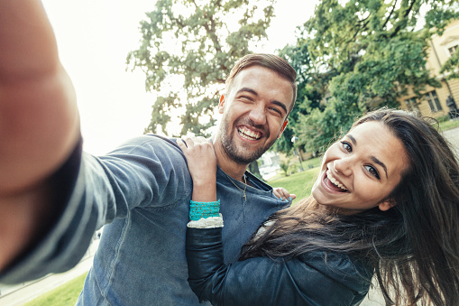 Young overjoyed couple making selfies in the park; hugging and laughing at camera.
