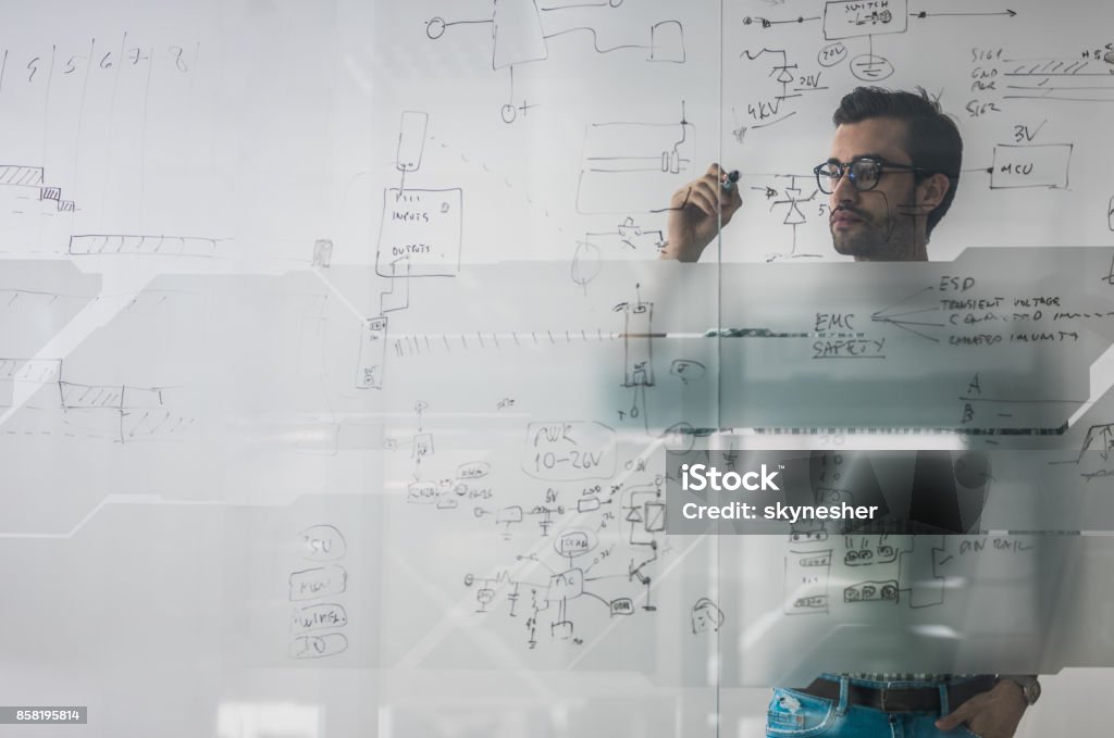 Male engineer working on new ideas and writing diagram on glass wall. Young engineer drawing a diagram on a glass in the office. The view is through glass. Complexity Stock Photo