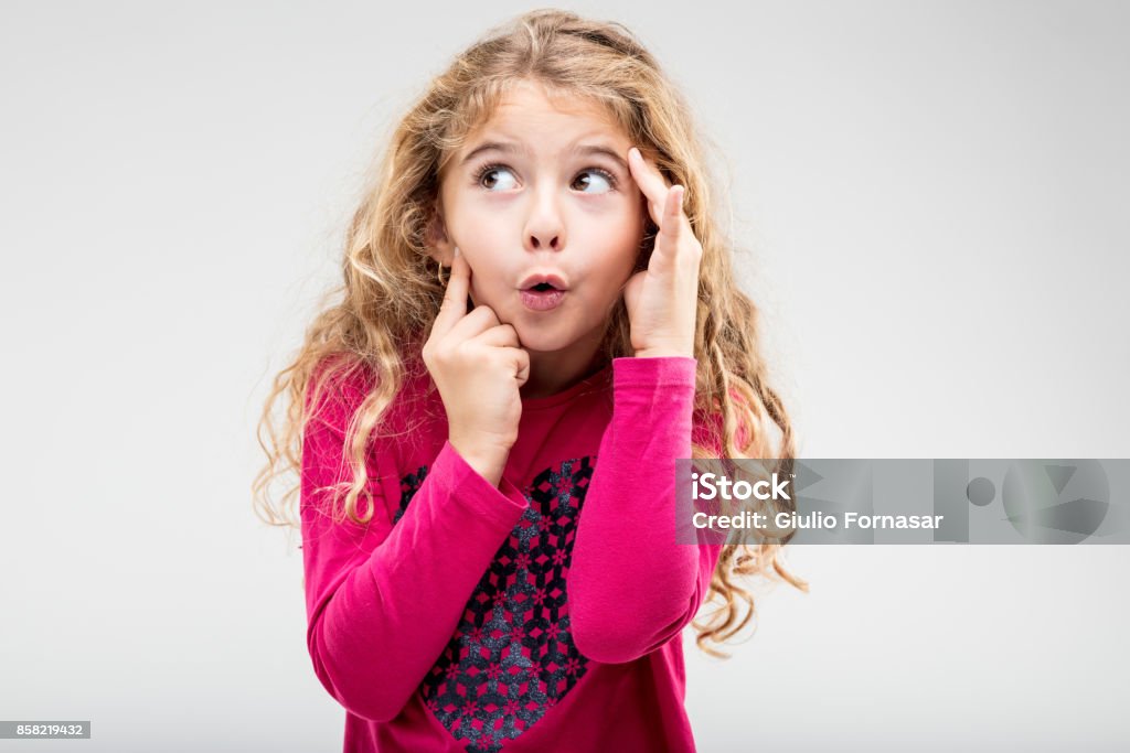 Fun playful little girl with a teasing expression Fun playful little blond girl with a teasing expression exclaiming Ooh holding her hands to her head as she glances up to the side Child Stock Photo