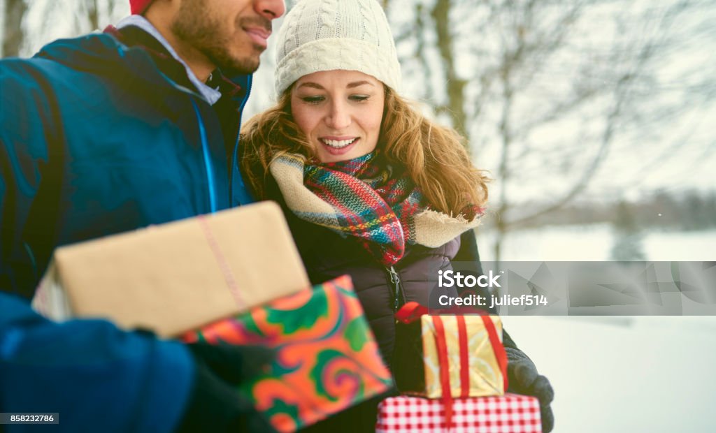 Smiling diverse couple holding Christmas presents while walking through a winter forest Happy multi-ethnic friends of caucasian and indian ethnicity carrying Chrismas gifts and walking in a snow covered park during holidays Shopping Stock Photo