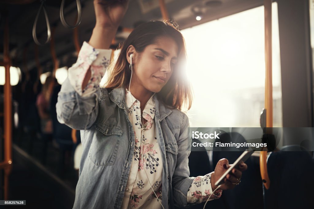 Young woman standing on a bus listening to music Young woman smiling while riding on a bus listening to music on a smartphone Commuter Stock Photo