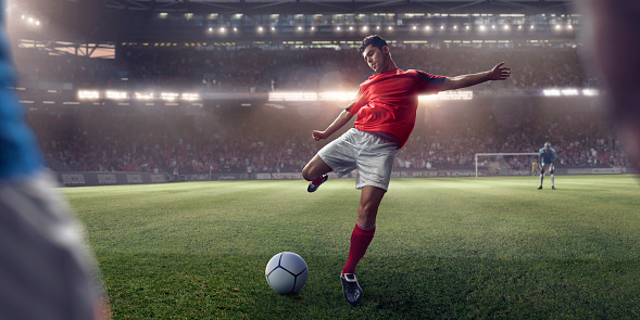 A shallow depth of field, close up view of a professional footballer in mid pose with right leg back and arms out, about to take a free kick as viewed from just behind an opposition player. The soccer player is playing a evening football match in a generic floodlit stadium full of spectators.