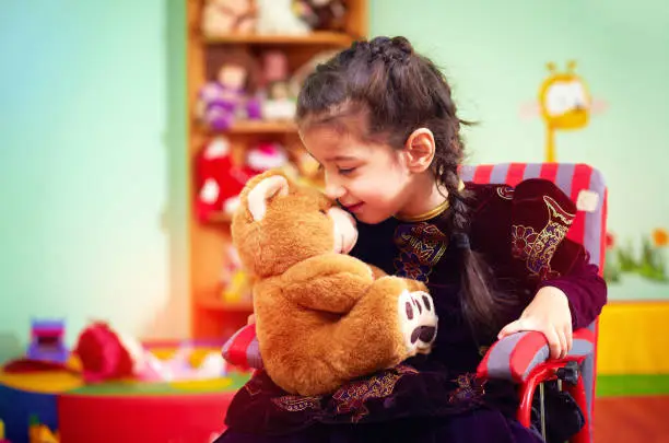 cute little girl in wheelchair telling her secret to plush bear in kindergarten for kids with special needs
