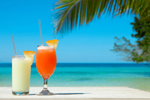 Two Exotic Cocktails At The Beach Two glasses of exotic cocktails on a white wood table with a caribbean seascape background. tropical drink stock pictures, royalty-free photos & images
