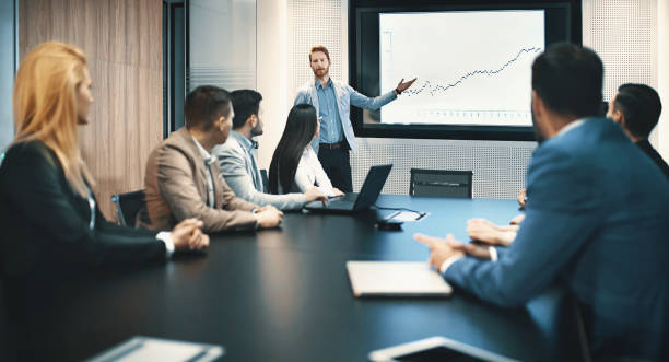 Board room meeting. Closeup of a board room meeting at a business company, usual scene at any modern company. There are five men and two women, one of the men is speaking in front of a large screen that's showing monthly revenue of their company. revenue stock pictures, royalty-free photos & images