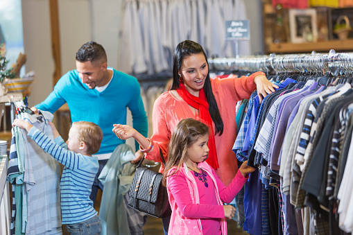 A mother and daughter shopping in a clothing store, looking at shirts hanging on a rack. Father and son are out of focus behind them.